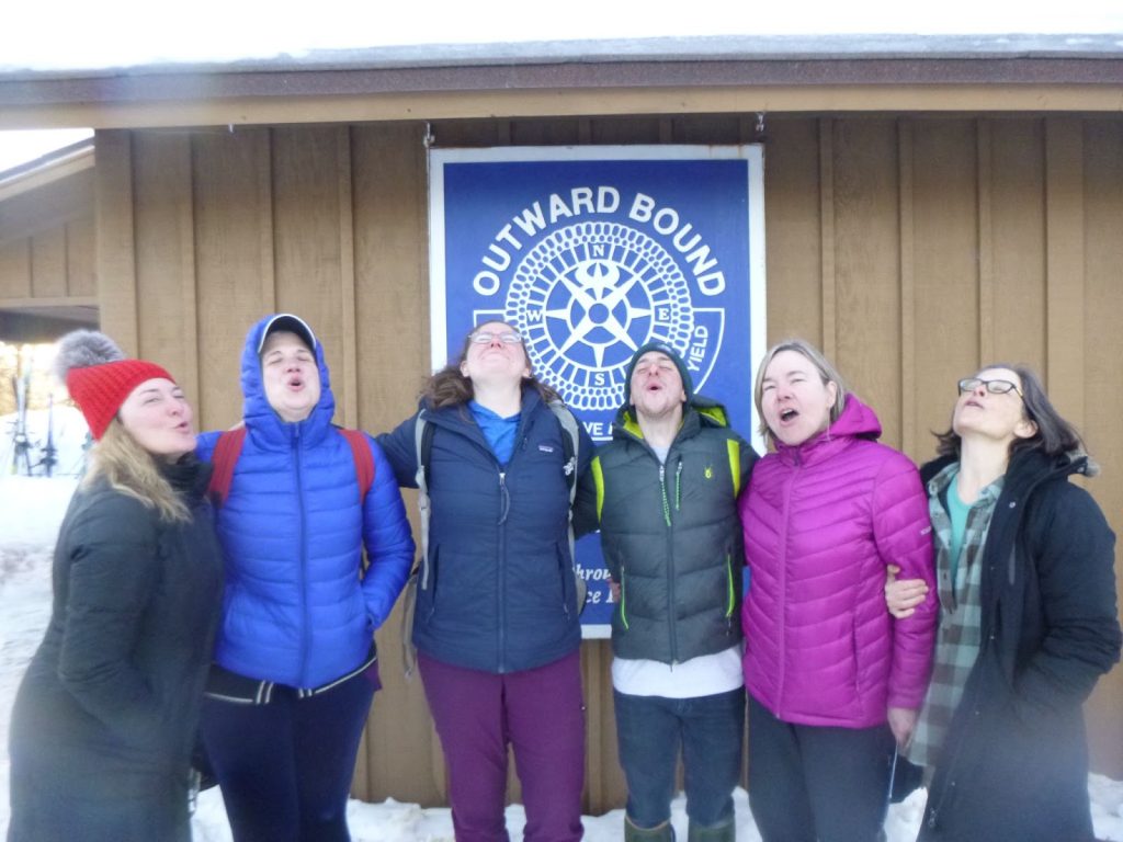 Six people pictured in winter setting howling towards the sky. Outward Bound logo behind them.